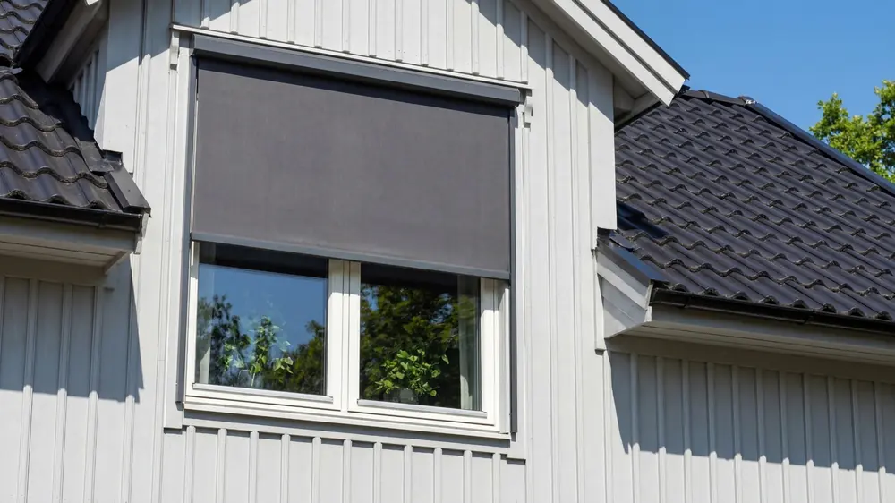 Grå vertikalmarkis nedfälld framför den övre delen av ett fönster på ett vitt trähus